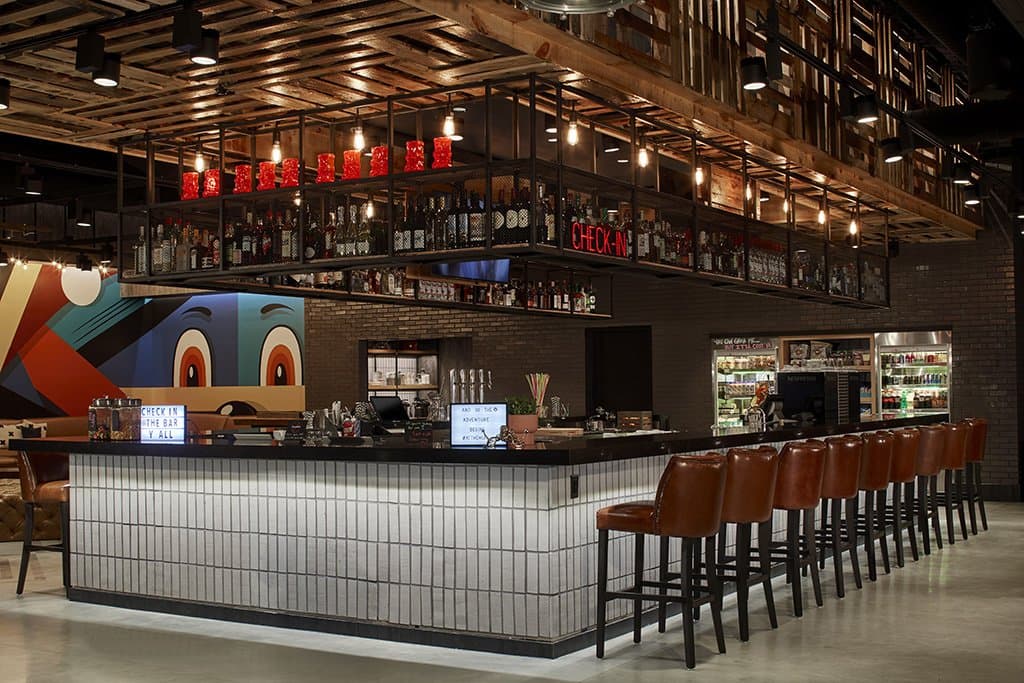 A subway tiled bar lined with leather wrapped barstools sits in the middle of a wide open room. Metal shelving hanging from the ceiling contains a variety of bottles.