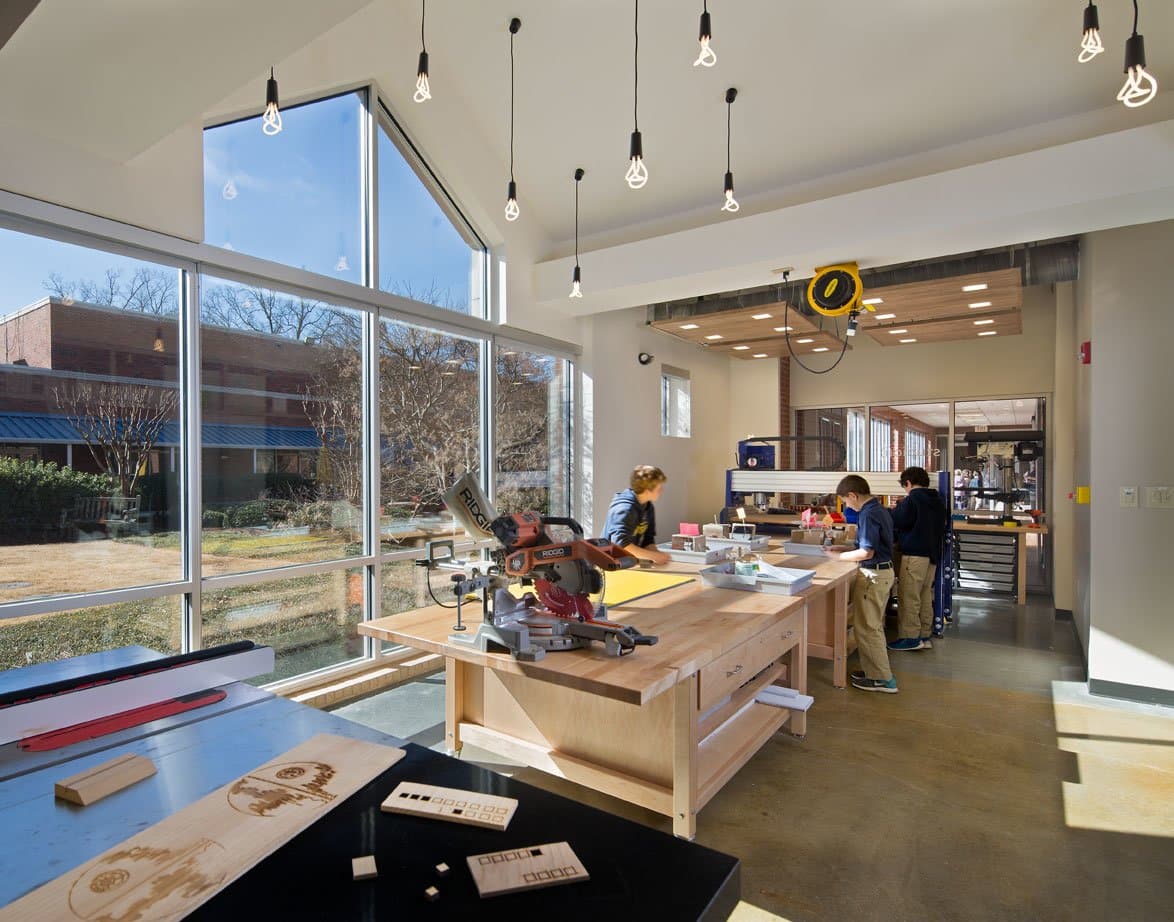 Children seen working in a woodshop with large windows, wood workbenches, and a RIGID brand miter saw.