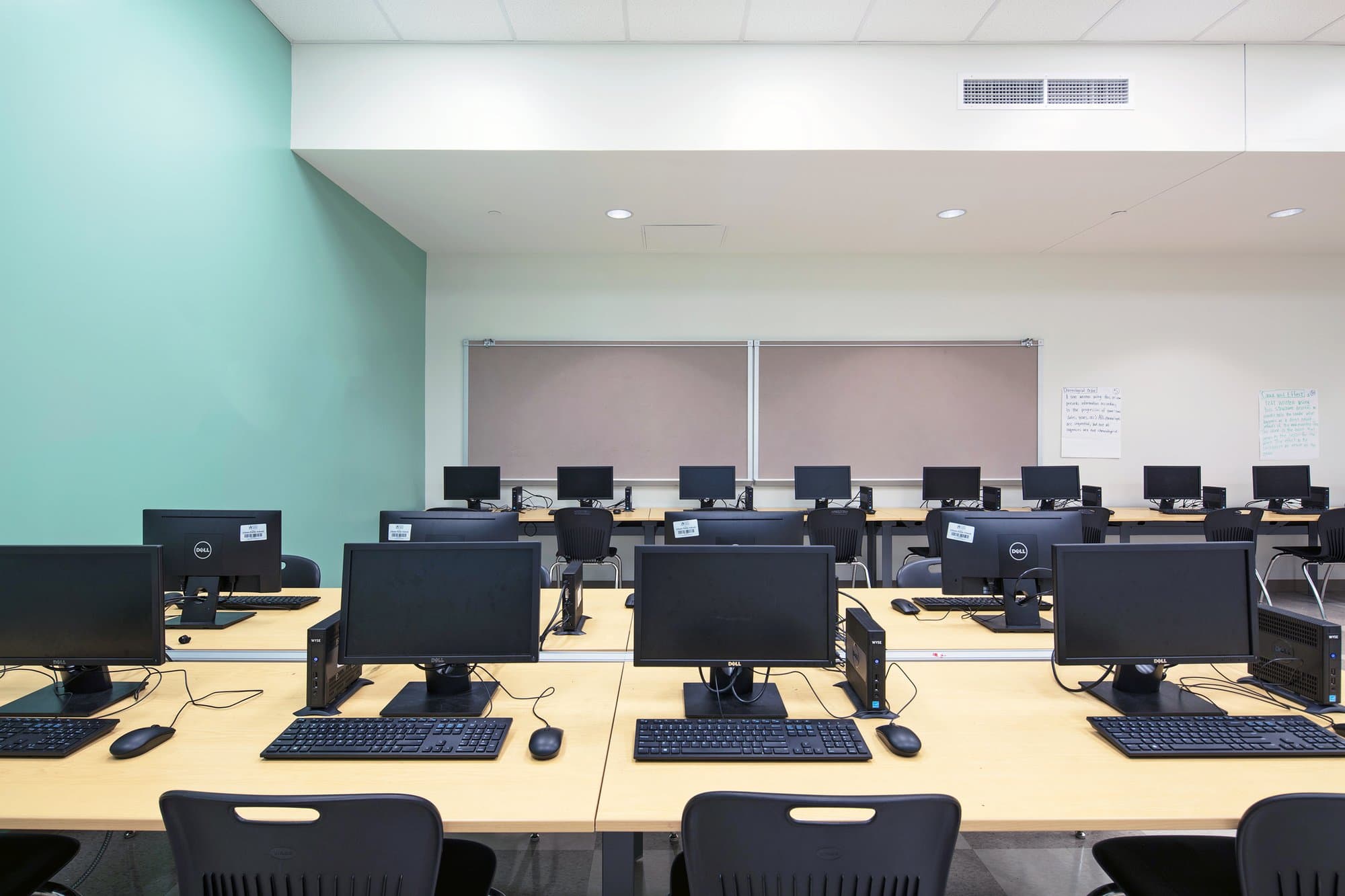 Computers and screens dot the light wooden surfaces of a computer lab.