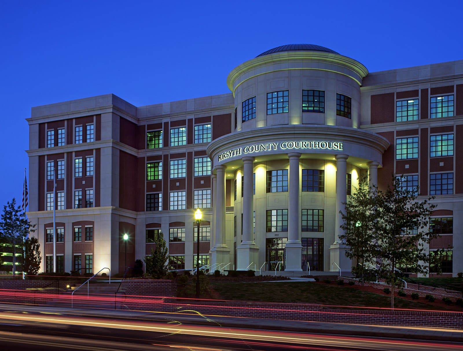 Picture taken of courthouse in dusk with car headlights and tail lights passing by.
