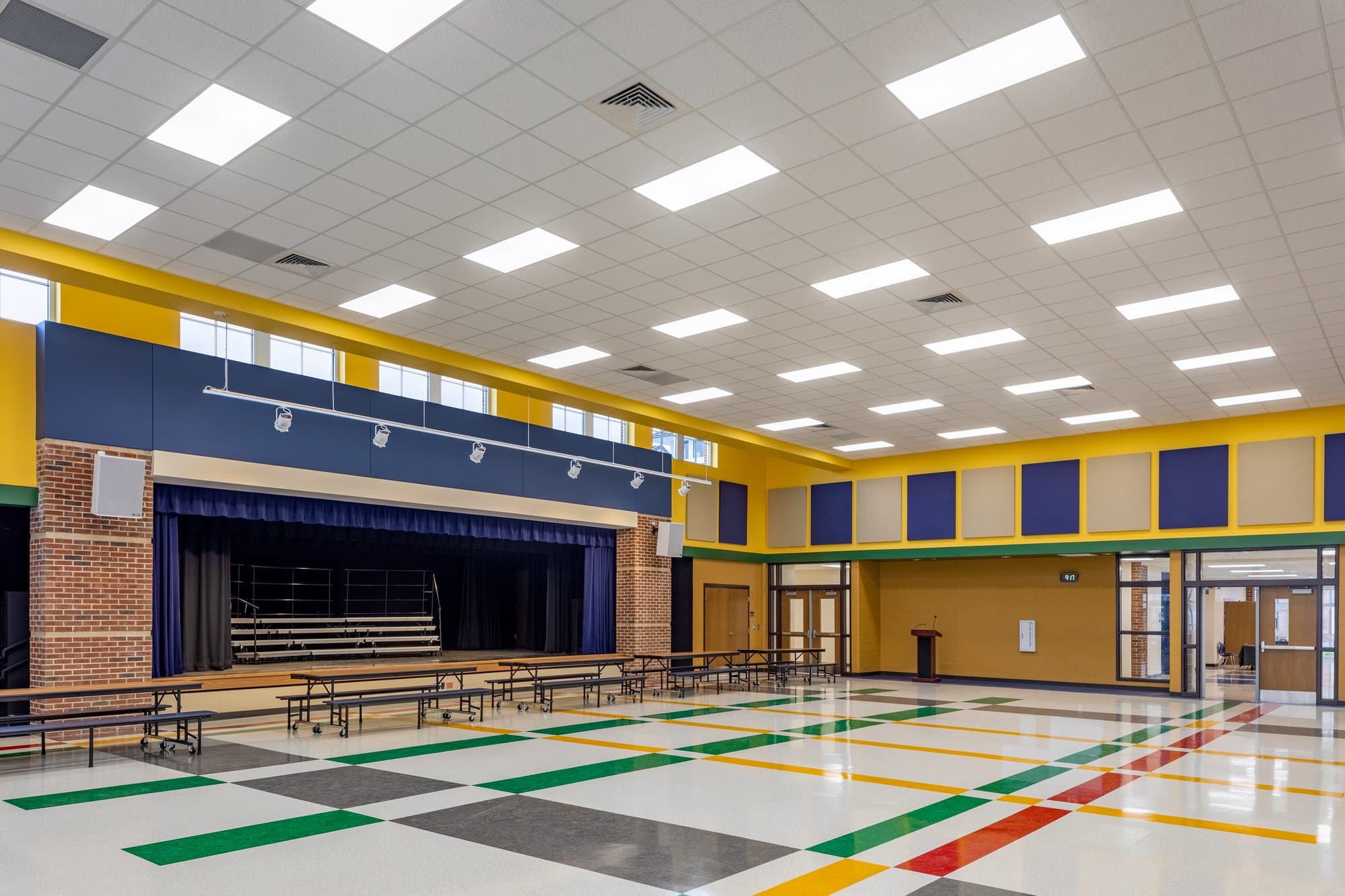 A multi-color tile floor dots a large open cafeteria/auditorium with a curtained stage off to the side.