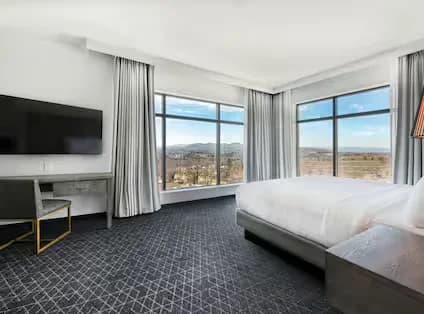 A spacious guest suite with dark colored textured carpet, neutral bed linens and curtains. Desk with chair and hanging TV. Suite has two windows at the corner of the room to maximize the views.