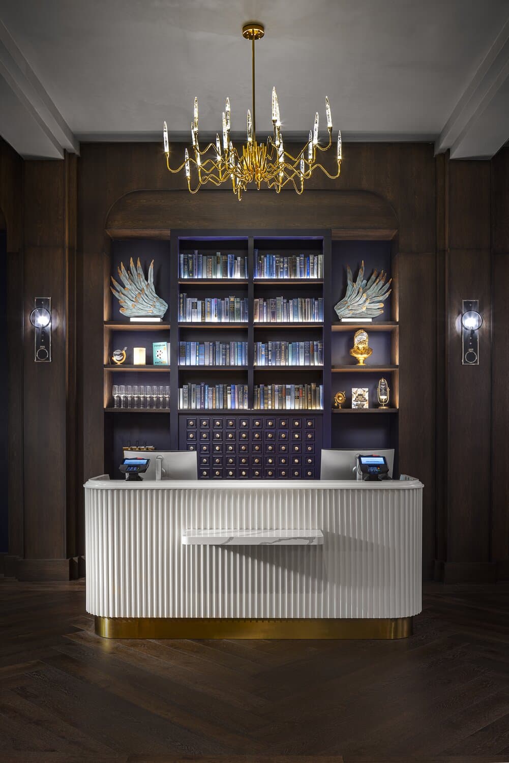 Guest services desk with white standing counter in front of a bookcase filled with books and side light sconces on either side.