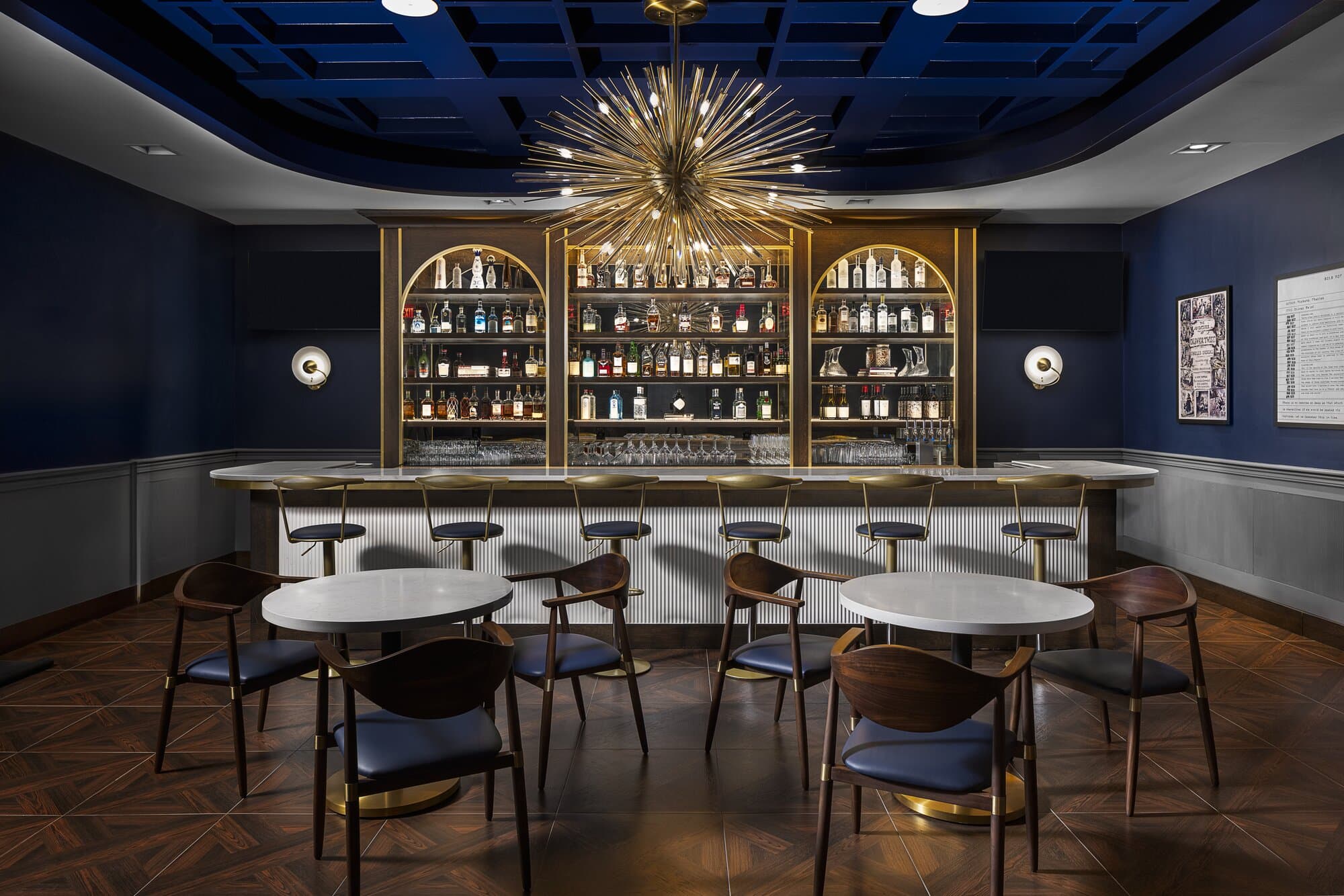 Intimate bar area with a navy blue ceiling and blue and white walls. A starburst chandalier above the bar with bar stools, tables and chairs.