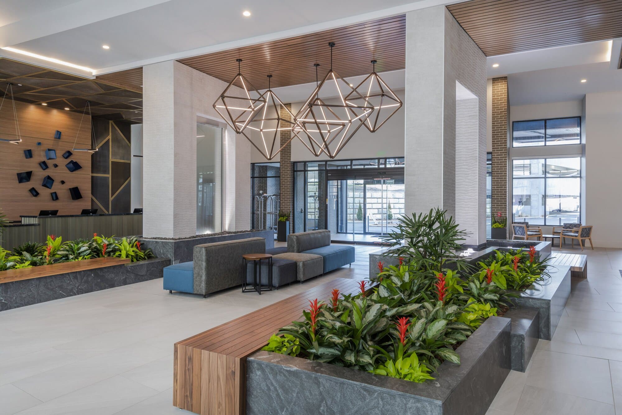 Foyer area, next to guest services showing four diamond shaped chandeliers at different sizes and heights grouped together. Walls with built in waterfalls stand on both sides of chandaliers surrounded by wood seating, lights, and foliage.